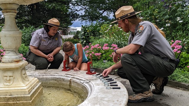 Two park rangers and a young child look into a microscope next to an outdoor fountain