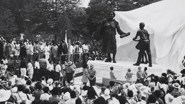 Large crowd of people gather outside in front of a large statue.