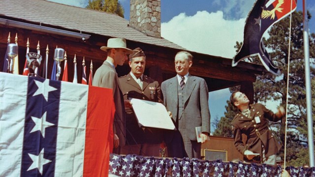 A black and white image of three men in suits sitting standing on a stage draped with a flag. 