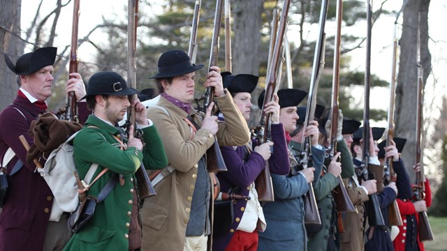 colonial reenactors, dressed in non-uniform clothing, perform drill with their muskets
