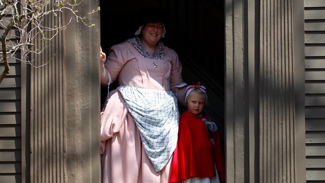 woman and young girl dressed in colonial clothing stand in the doorway of a brown colonial house