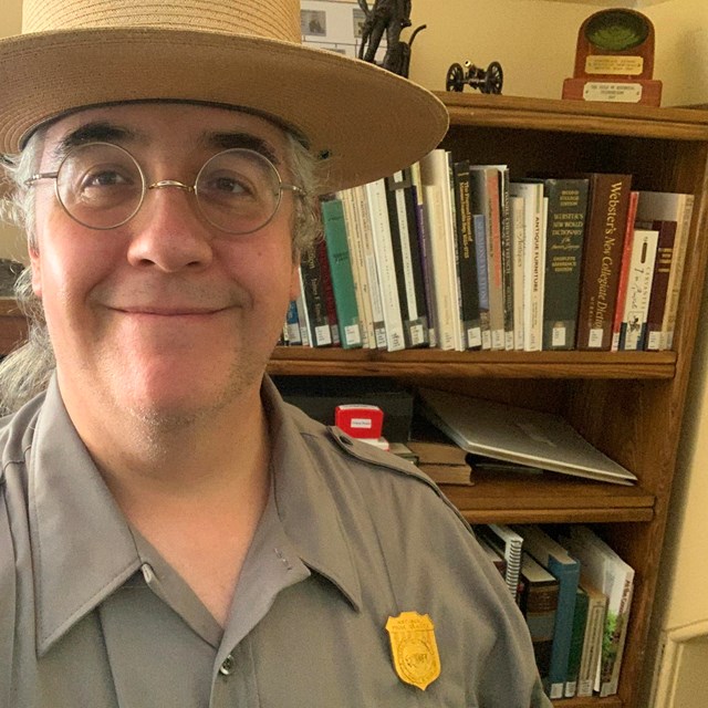 Man in NPS uniform and flat hat stands in front of a book shelf