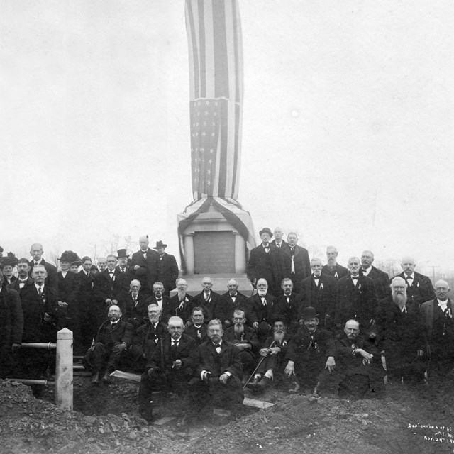 Black and white photo of old white men in dark suits surrounding a flag draped monument.