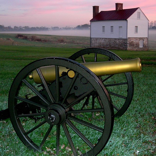 A Civil War cannon in front of a small two story house with the sun setting behind it.
