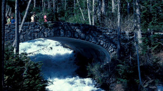 A stone-faced bridge over a raging white river in a forest. 