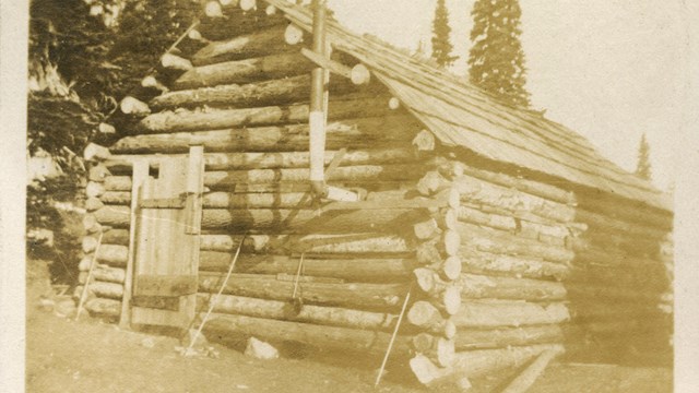 A roughly constructed wood cabin with a single door and no windows with a metal stove pipe. 