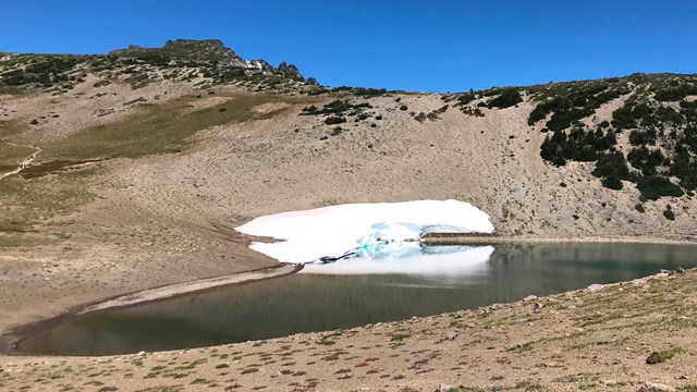 A small lake next to a rocky ridge with a small snowbank clinging to one shore.