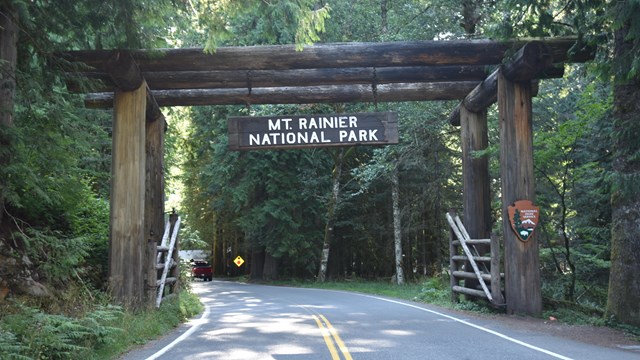A paved road bordered by forest enters a log entrance arch with a Mt Rainier National Park sign.