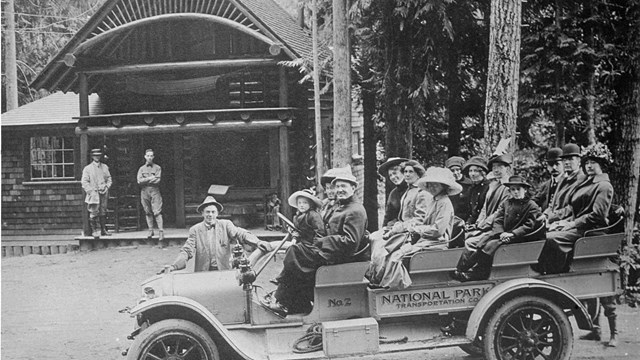 Historic photo of visitors sitting in an open-top vehicle parked in front of a small wood building. 