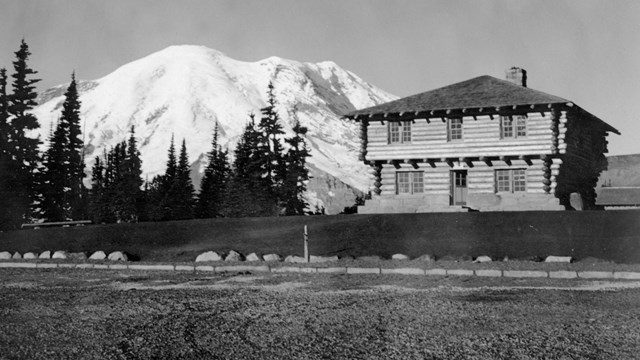 A two-story square log building and large dirt parking lot in front of a glaciated mountain. 