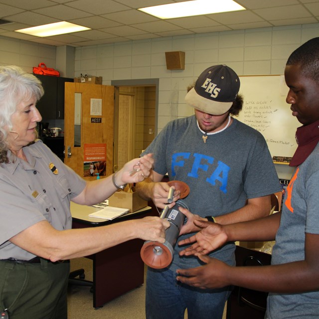 Two high school students examining a grey cylinder with a ranger. 