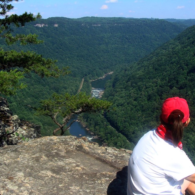 A woman sitting on a rocky outcropping overlooking a deep green gorge with a river at the bottom