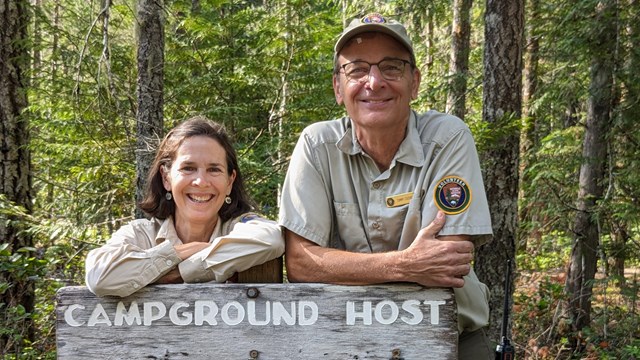 Two smiling volunteers with a sign that reads Campground Host with forest behind them.