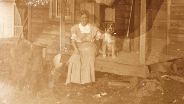 A old sepia photo of a woman in long dress sitting on the wooden porch of a cabin with a dog