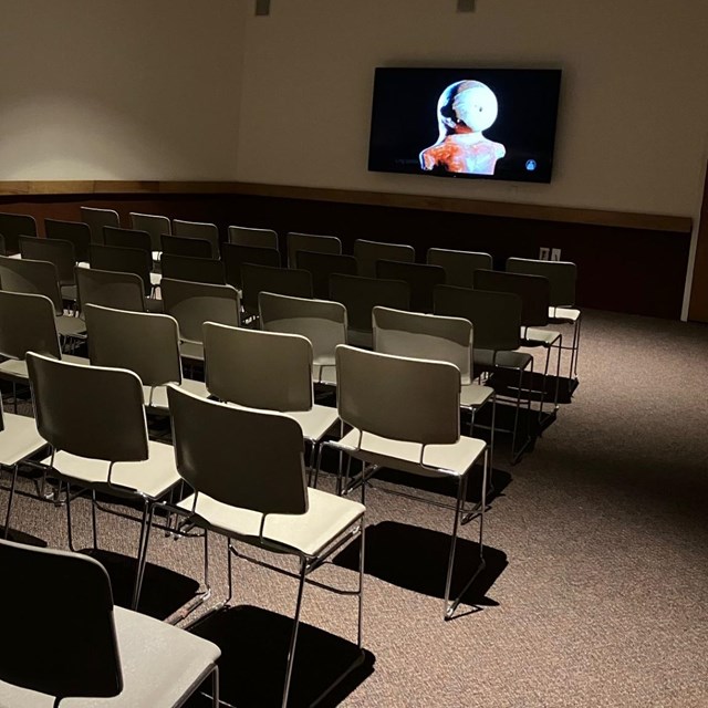 NPS Photo of the Visitor Center Theater, with several rows of chairs facing a mounted TV showing the