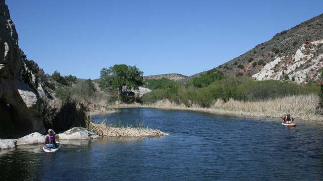 People sitting on paddle boards make their way down a river, set in a semi-arid canyon.