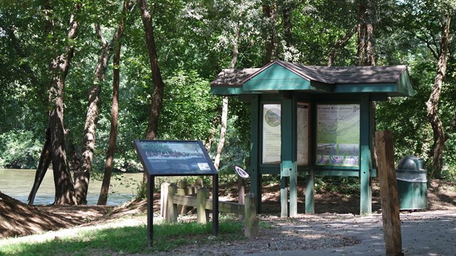 A sign kiosk beside a paved trail in the shade of trees