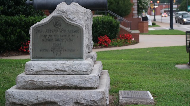 A stone marker with small black cannon in front of a brick building and beside a sidewalk