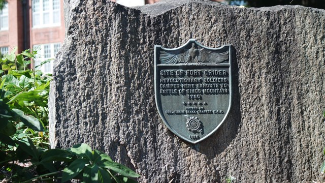 A metal plaque mounted on a stone marker in front of a brick building