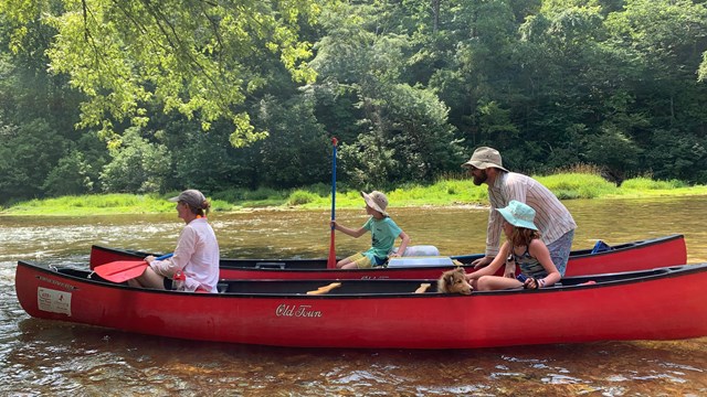 Two red canoes float in shallow water. There are two adults, two kids, and a dog.