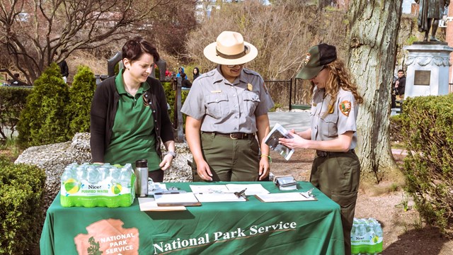 Two park rangers & a green-clad volunteer stand at a National Park Service info table