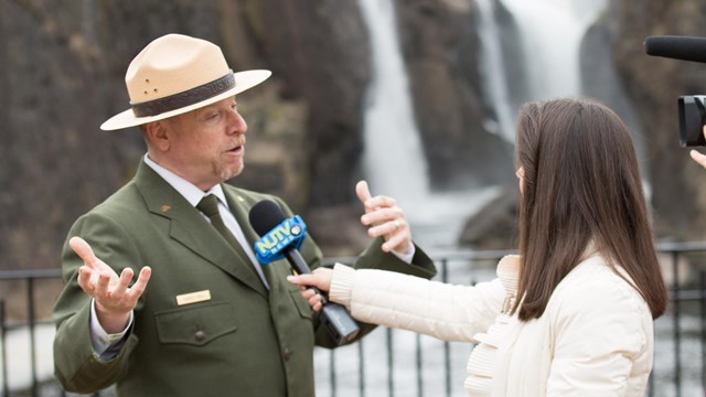 A reporter holds a microphone towards a park ranger in dress uniform in front of a waterfall