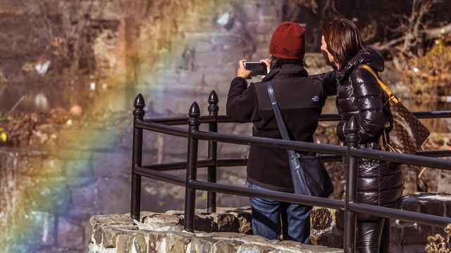 Two women take a photo of a bright rainbow at a waterfall overlook railing