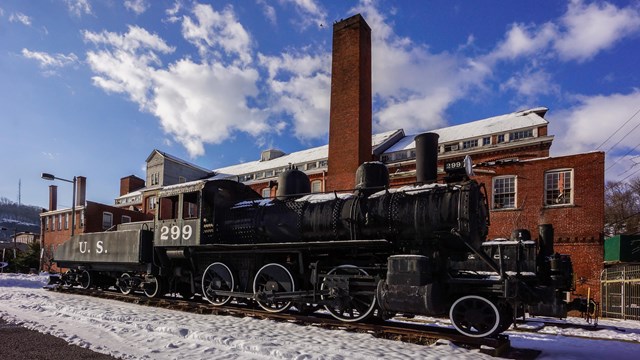 A black 2-6-0 steam locomotive with tender sits in the snow outside the brick Paterson Museum