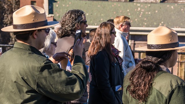Visitors & park rangers look towards something at right - one holds a phone up to record it