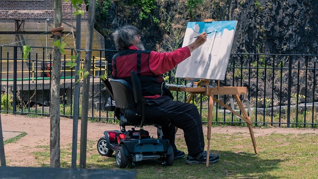 A man in a power wheelchair paints a view of the Great Falls with an easel