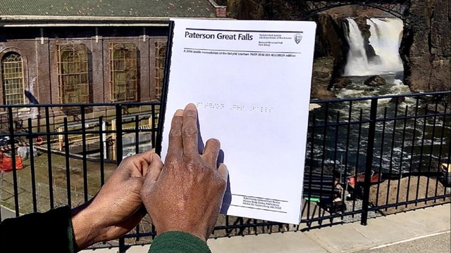 A hand runs fingers over a braille brochure for Paterson Great Falls, a waterfall in the background