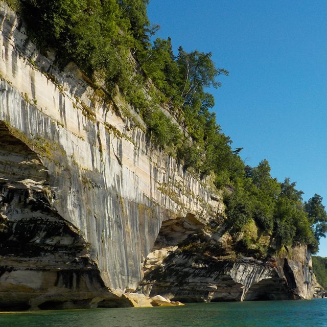 Tree-topped black streaked cliff wall with eroded out portions at the Lake Superior waterline.