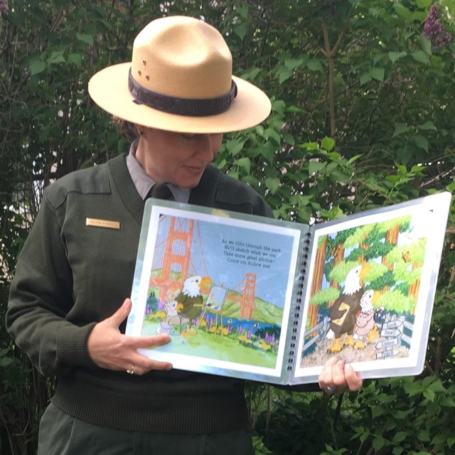 Park Ranger standing outside with an open book facing everyone as he reads the story.