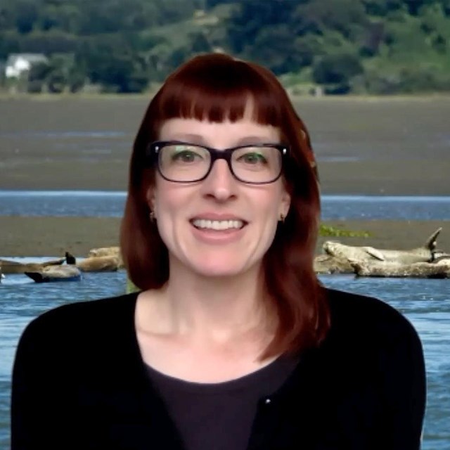 A smiling, red-headed woman wearing a black shirt appears in front of harbor seals on a sand bar.