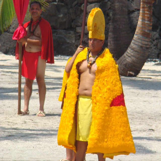 Man in feathered cloak and helmet of the aliʻi (royalty) walks through the Royal Grounds