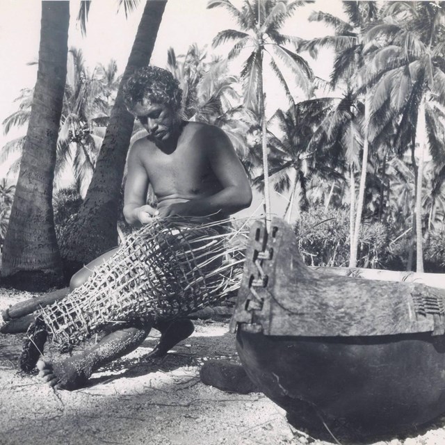 Man in traditional clothing weaves a fishtrap while sitting under coconut trees in the Royal Grounds