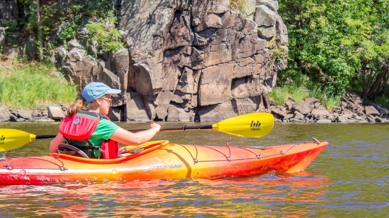 A kayaker paddles on a river with rocky bluffs.