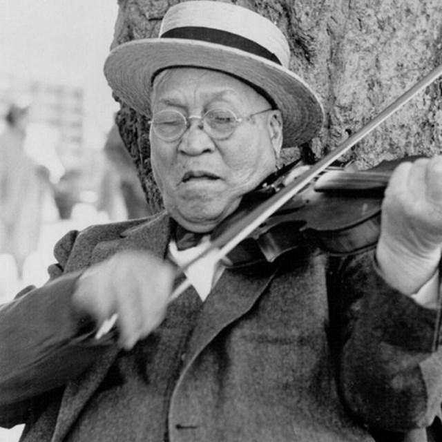 A man in a three piece suit and a straw boater vigorously plays a fiddle