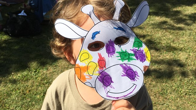 Child holds a colored paper animal mask over their face.