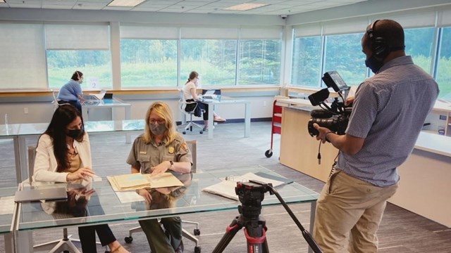 A ranger sits with a researcher reviewing documents while being filmed.