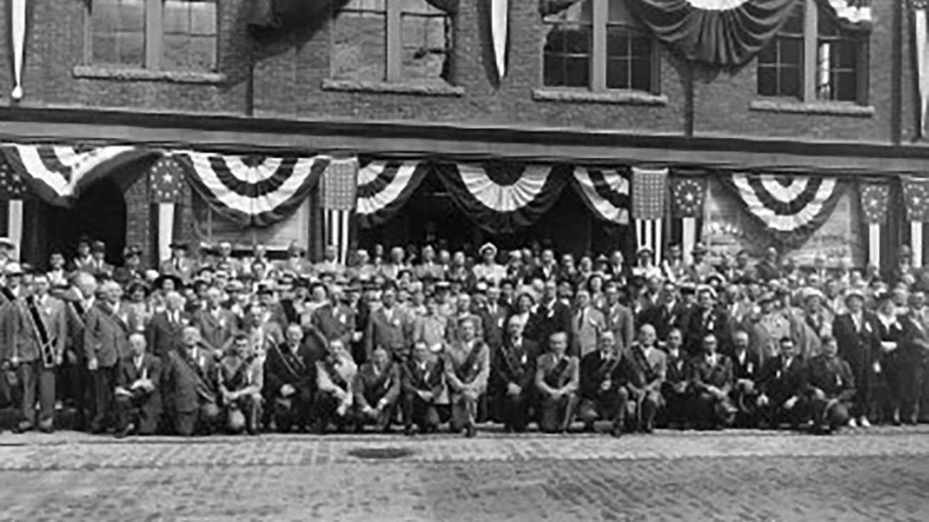 A large group of people gather in front of a three-story brick building.