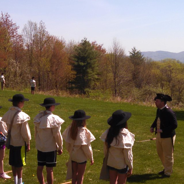 Students stand on a grassy hill looking at a park ranger dressed in 18th century clothing. 