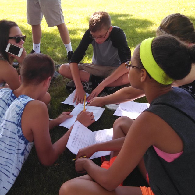 Students sit in a circle formation on the grass while working on educational work sheets.