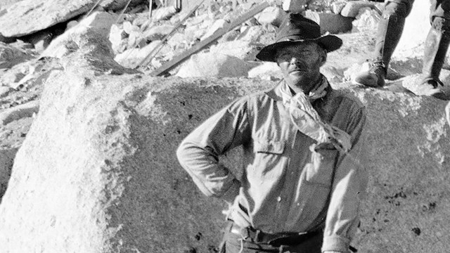 A man with a brimmed hat, white scarf and buttoned shirt, with one arm akimbo in front of a big rock