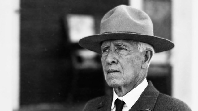 An older man wears a wide and flat-brimmed hat with a suit and tie. He gazes to the left..