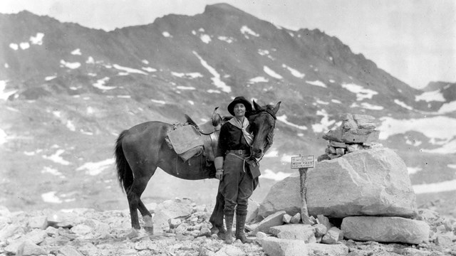 A woman stands with a horse next to a sign and a large boulder, surrounded by snowy mountains.