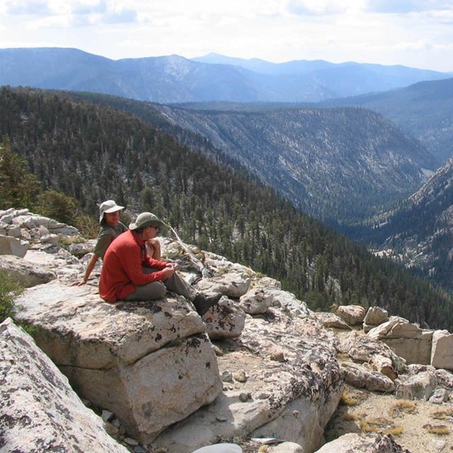 Two people sit on a boulder looking out across a wide forested valley and distant mountains