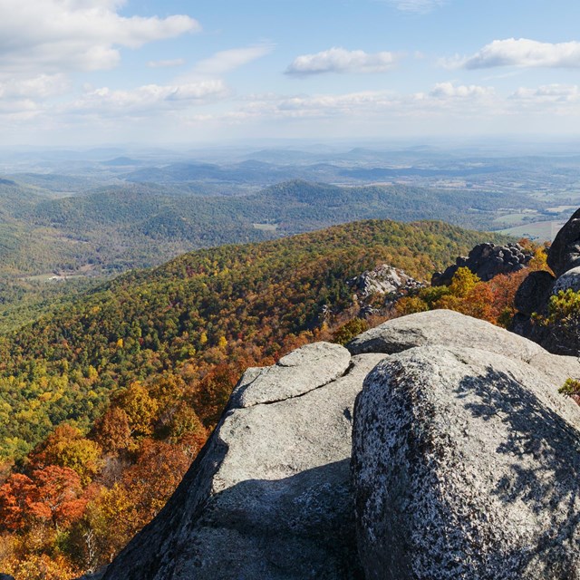 Large rocks stand in the foreground of a view overlooking trees displaying fall color in the valley