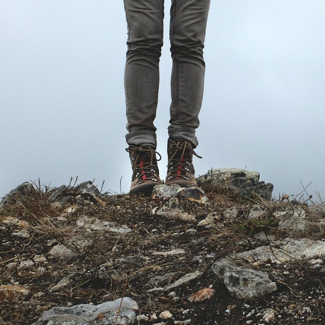 A close-up of a hiker's legs with hiking boots, standing on a rocky viewpoint.