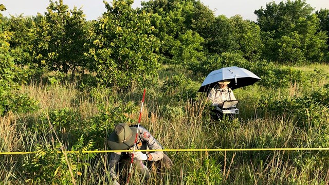 Two field crew members in a grassy field, one observing, the other taking notes.